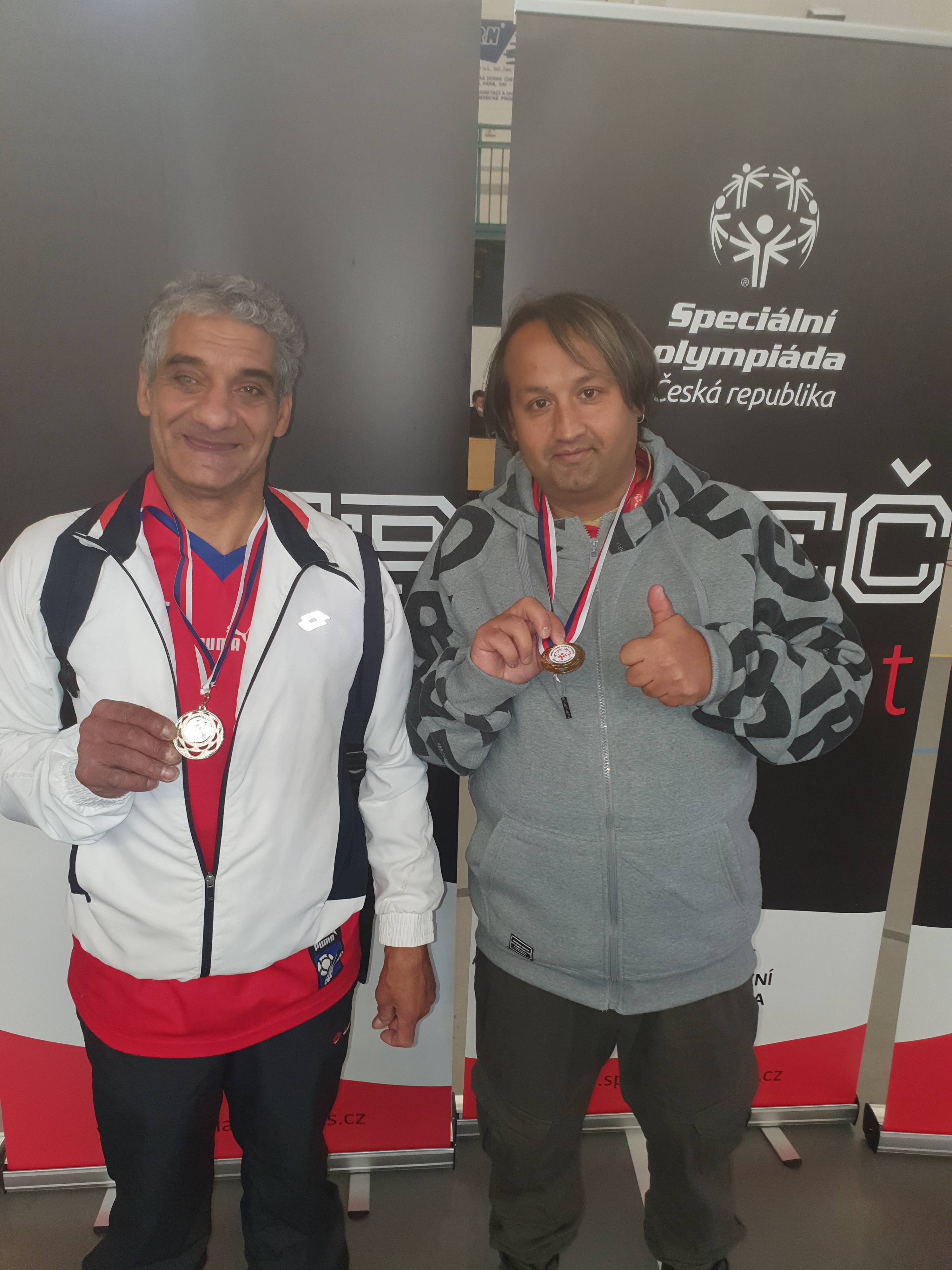 dvě usmívající se muži stojící před plakáty s logem Speciální olympiády mající kolem krků medaile, muž vpravo ukazuje zdvižený palec