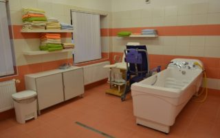 koupelna s oranžovým obložením s masážní vanou, kolečkovým křeslem, vyvýšeným křeslem, skříňkou a policemi s ručníky