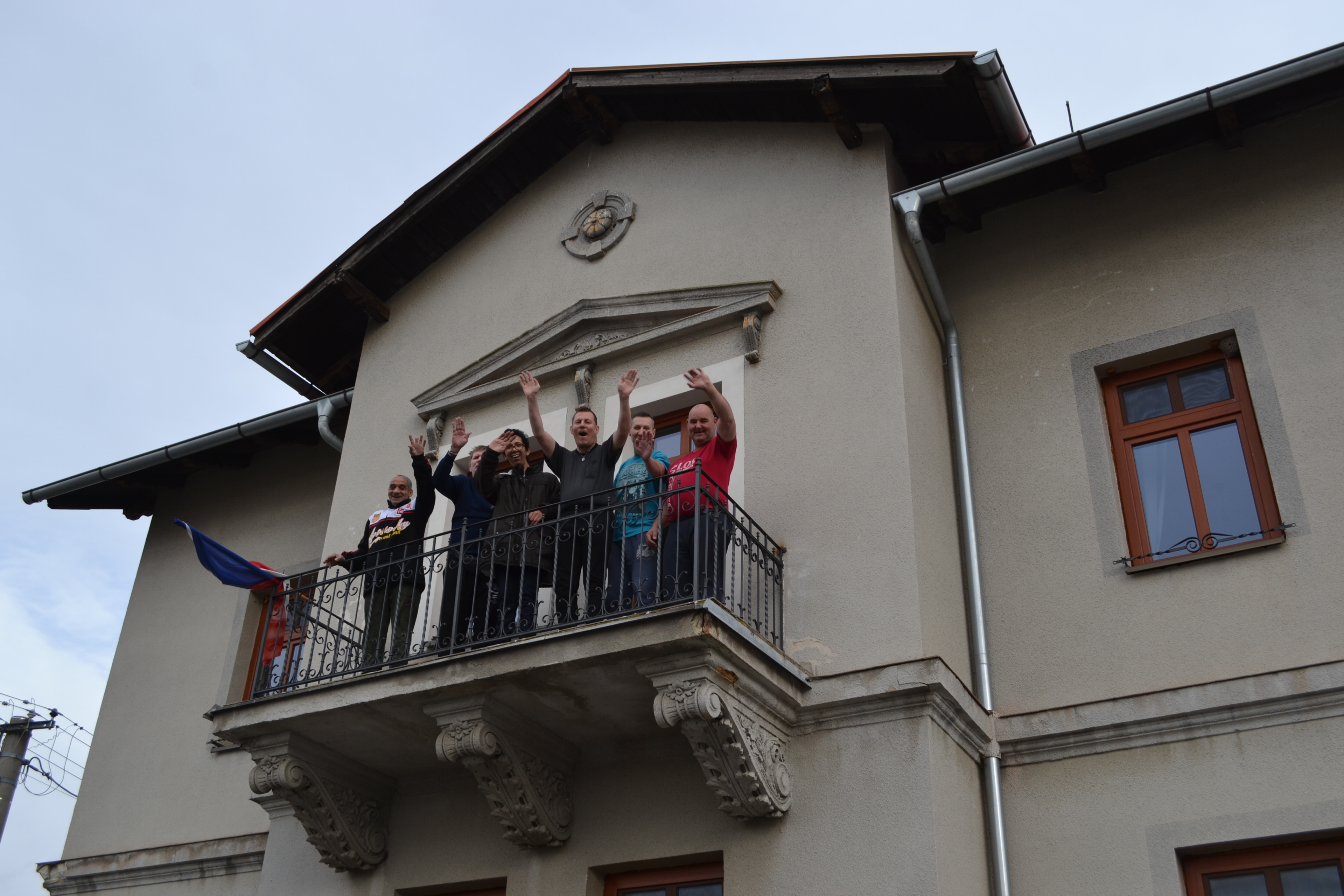 jásající skupina osob focena zespodu, stojící na balkoně šedého domu s českou vlajkou
