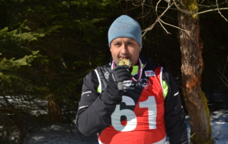 muž ve sportovním zimním oblečení stojící v zasněženém lese, kousající do zlaté medaile