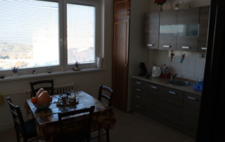 kuchyně s kuchyňským koutem, velkým oknem a čtvercovým jídelním stolem obestavěným čtyřmi dřevěnými židlemi