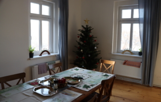 místnost s bílými stěnami, v rohu vánoční stromek, v popředí dlouhý dřevěný stůl obestavěný dřevěnými židlemi v rustikálním stylu
