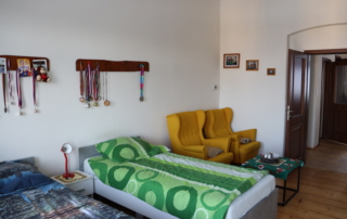 pohled na dvě postele, v rohu dva žluté ušáky se stolečkem, na stěně věšáčky s množstvím medailí