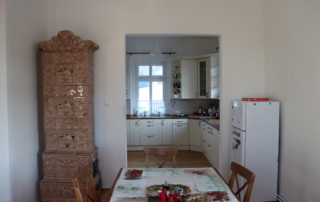 vánočně prostřený dřevěný stůl s židlemi v místnosti s kamny a lednicí, v pozadí kuchyně v rustikálním stylu