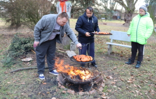 tři muži stojící kolem ohniště, věnující se opékání špekáčků na roštu