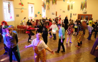 skupina osob tančící v kostýmech v prostorném sále s dřevěnou podlahou, zdi vyzdobené balonky a jinými dekoracemi