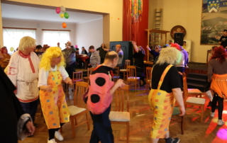 skupina osob tančící v kostýmech v prostorném sále s dřevěnou podlahou, zdi vyzdobené balonky a jinými dekoracemi