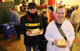 dvě usmívající osoby v kostýmech držící kus dortu na papírovém tácku, v pozadí tancující osoby